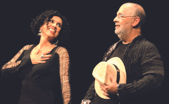 Imagem mostra uma mulher e um homem, de mãos dadas, sorrindo em cima de palco