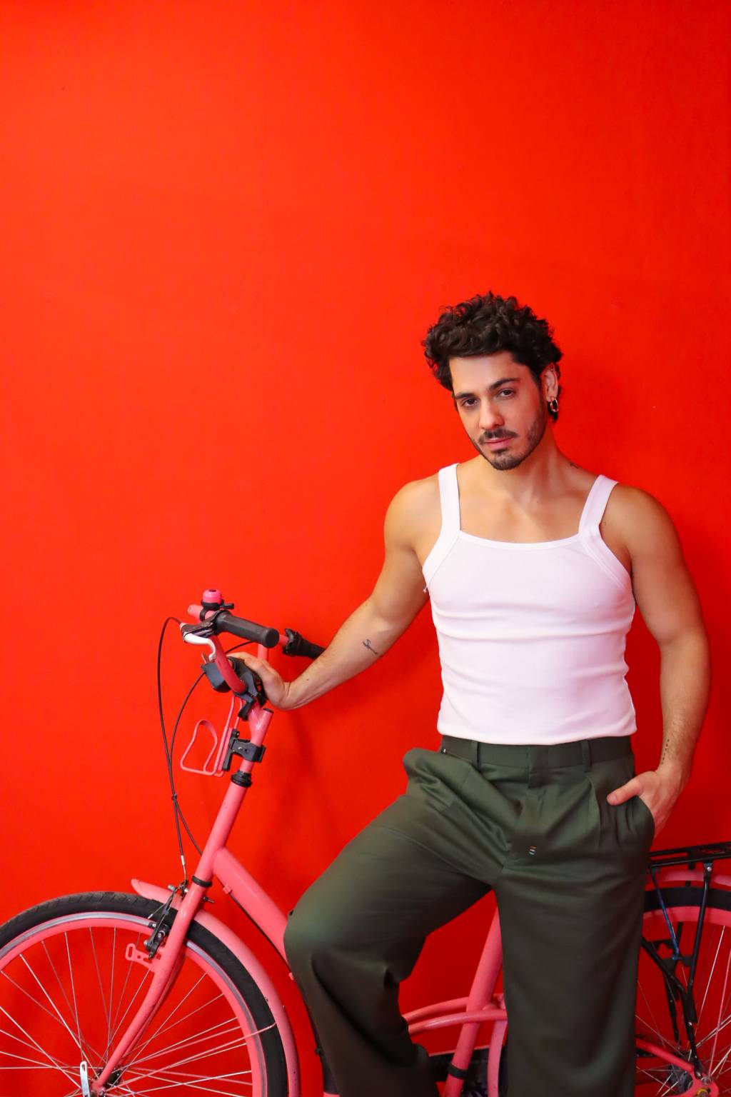 Diego posa sentado em bicicleta rosa e parede de fundo bem vermelho. Veste regata branca e calça escura.