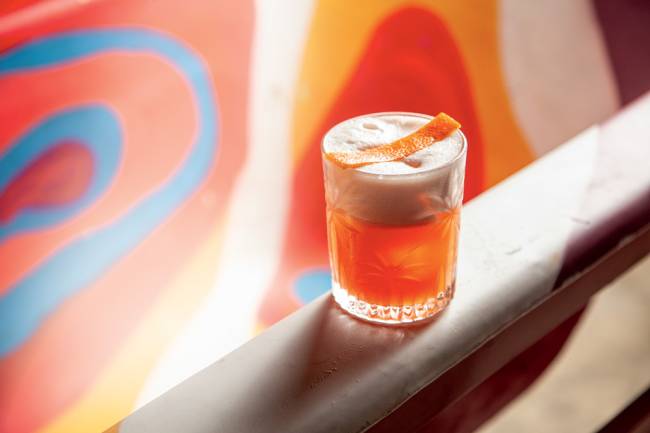 Drinque amaro mio_3.jpg Copo baixo com conteúdo laranja, dois dedos de espuma branca e tira de casca de laranja apoiada sobre ela, sobre uma espécie de parapeito claro com um fundo com estampa psicodélica
