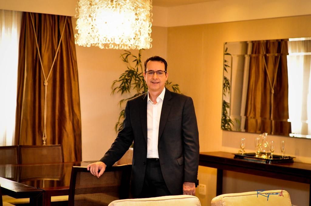 Elias Rodrigues, presidente da HCC Hotels, posa de terno em sala iluminada. Apoia uma das mãos em cadeira de madeira e aparece com lustre no alto em destaque. Espelho, cortina marrom e planta ao fundo.