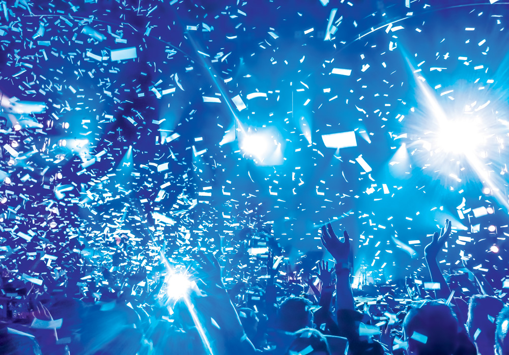 Imagem genérica de pessoas curtinho um show de música. É possível ver apenas seus braços para cima, cobertos por confetes e luz azul