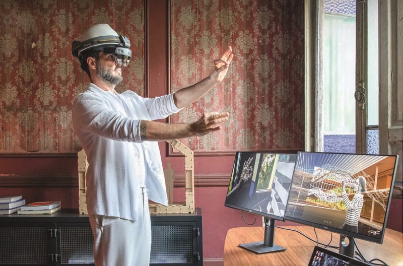 Guto Requena posa com as mãos erguidas em frente ao rosto enquanto visualiza imagens em um aparelho de realidade virtual. Veste camisa e calça brancas em escritório com computadores à direita e fundo estampado na cor vinho.