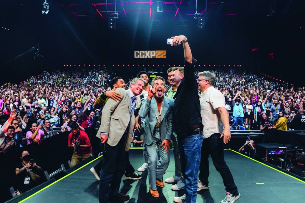 Imagem mostra grupo de homens em cima de palco, tirando selfie com uma multidão no teatro