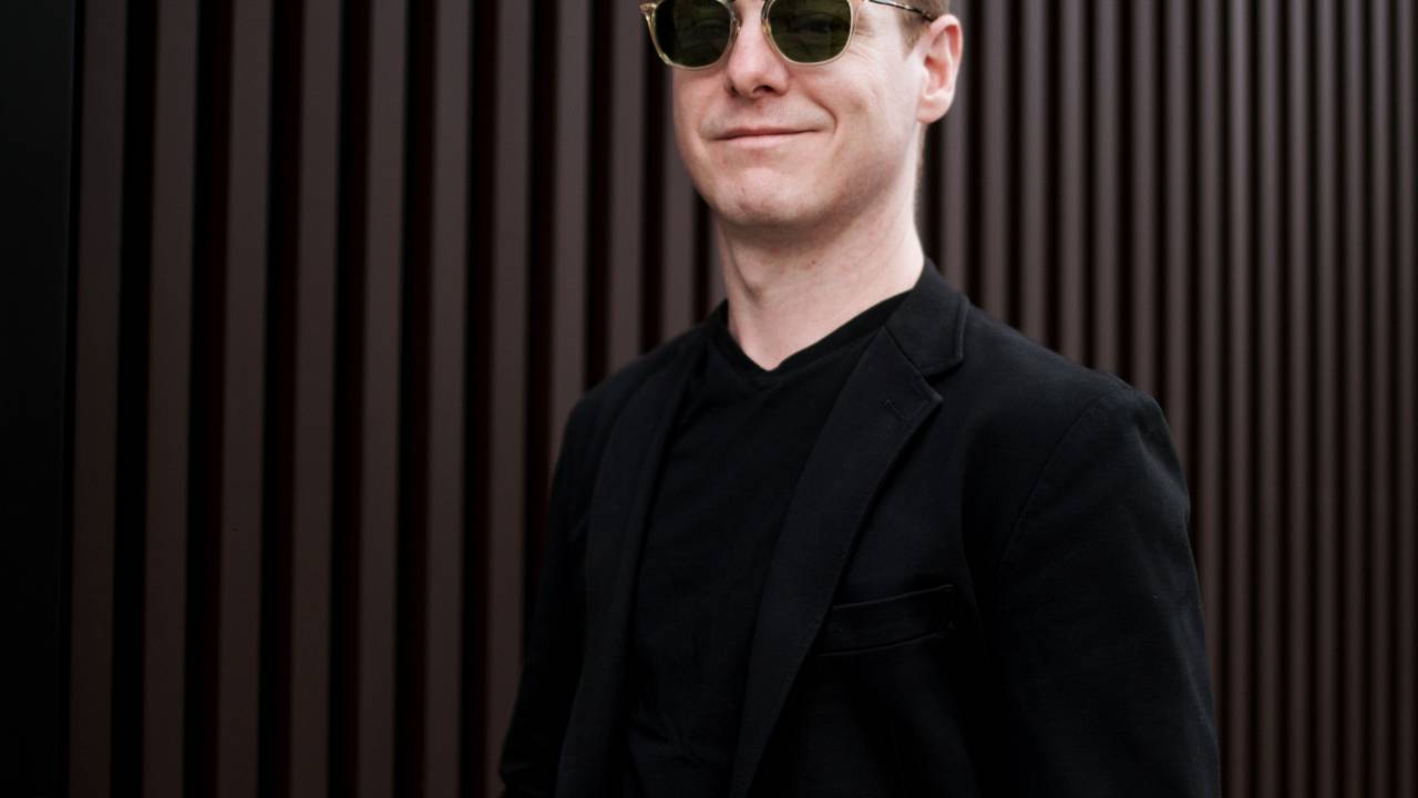 O designer Jader Almeida posa de óculos escuros, topete de cabelos escuros castanhos, roupas pretas e fundo em tom marrom.