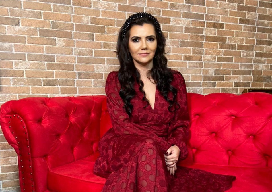 Espiritualista Kelida Marques posa sentada de pernas cruzadas em sofá vermelho. Veste vestido também vermelho e tem cabelos pretos longos.