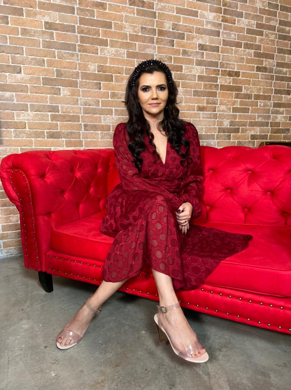 Espiritualista Kelida Marques posa sentada de pernas cruzadas em sofá vermelho. Veste vestido também vermelho e tem cabelos pretos longos.