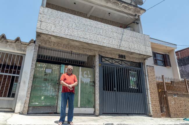 _MG_7371.jpg Foto de Adolfo, usando camiseta vermelha e calça jeans, em frente a sua casa