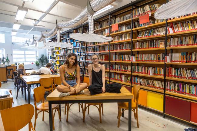 Jovens de 17 anos sentadas de pernas cruzadas sobre mesa com biblioteca ao fundo