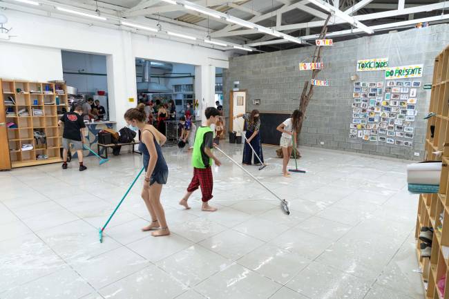 Alunos portando rodos lavam o páteo da escola