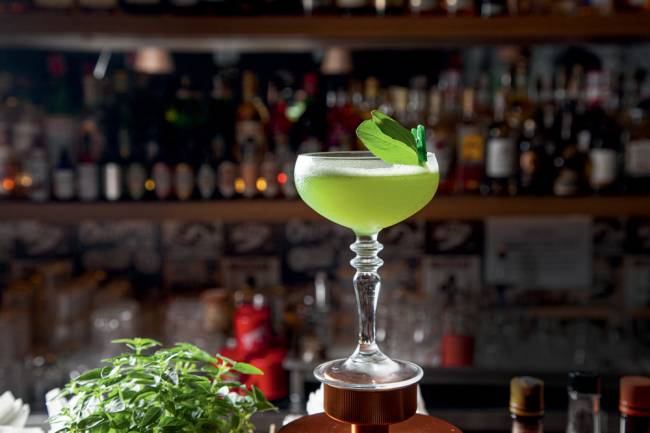 Taça alta arredondada contendo bebida verde translúcida com folha verde presa na borda com miniprendedor; ao fundo, as prateleiras de um bar
