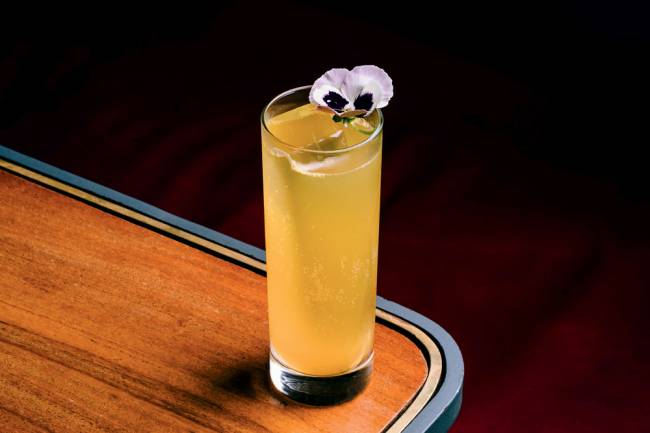 Passiflora Fizz SubAstor Copo alto com conteúdo amarelo translúcido e flor do tipo amor perfeito na borda, sobre mesa de madeira clara