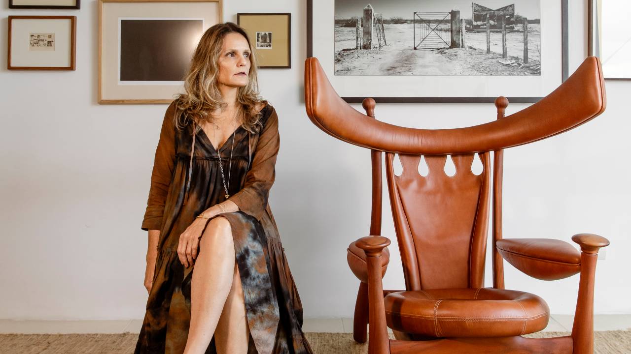 Renata Aragão posa sentada ao lado de cadeira com pontas avantajadas de madeira. Veste vestido todo preto, tem cabelos claros longos. Ao fundo, quadros em diversos tamanhos exibem paisagens.