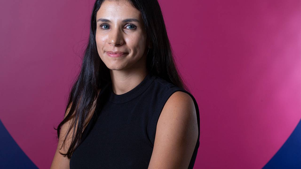 Talita Lacerda, CEO da Petlove, posa de braços cruzados com fundo que estampa coração rosa. Veste regata preta e tem cabelos pretos longos.