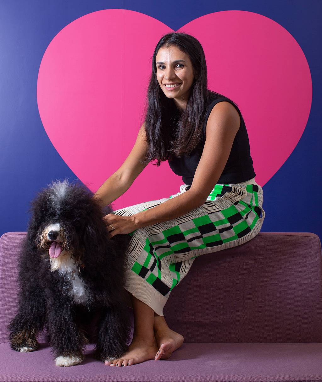 Talita Lacerda, CEO da Petlove, posa sentada com cachorro preto ao lado e fundo com coração rosa. Tem cabelos pretos longos e veste jeans clara.