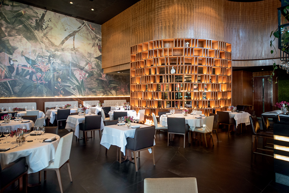 Salão do restaurante Urus exibe mesas vazias com toalhas brancas, cadeiras pretas e grande estante iluminada em madeira ao fundo.