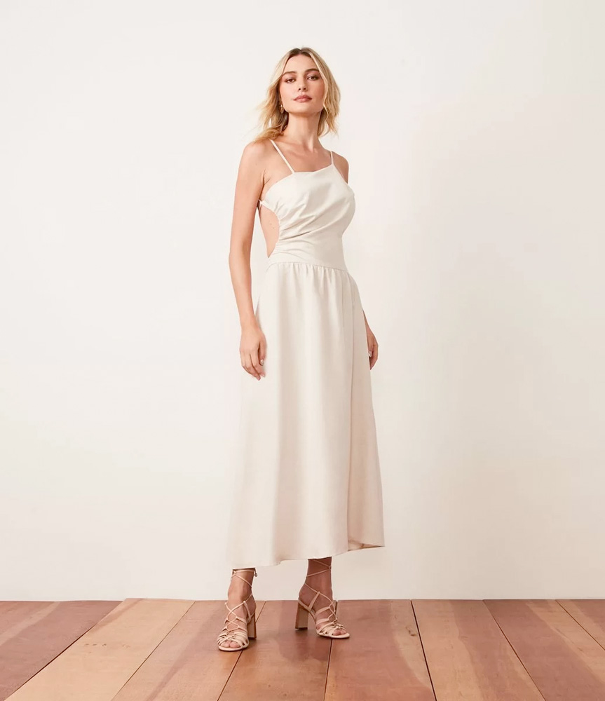 Modelo branca, magra e loira usa vestido off-white sem estampa. Ele tem al&ccedil;as de tiras, decote assim&eacute;trico, repuxo nas costas e comprimento midi