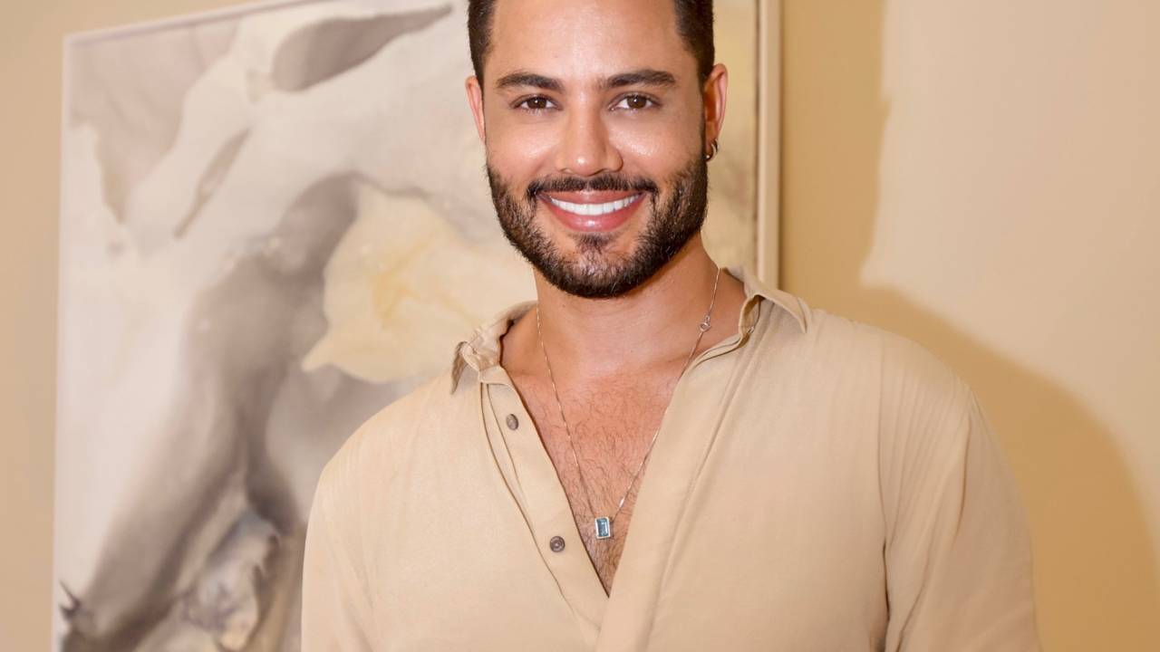 Hair stylist Well Dias sorri para a câmera. veste blusa bege com botões abertos, tem cabelos pretos e barba.