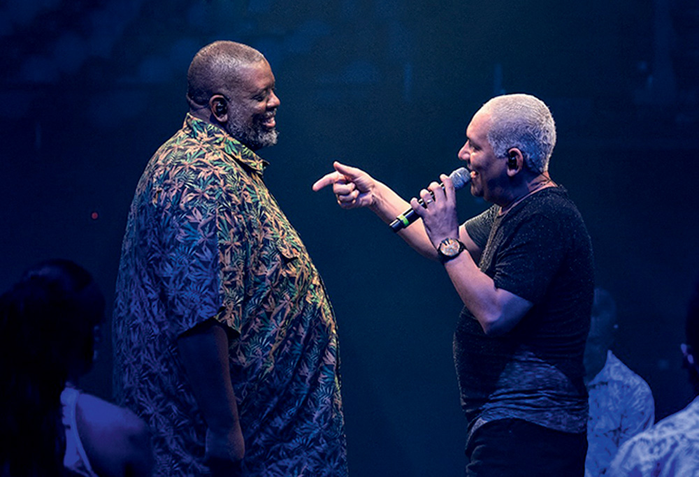 Imagem mostra dois homens em frente ao outro, um cantando em um microfone