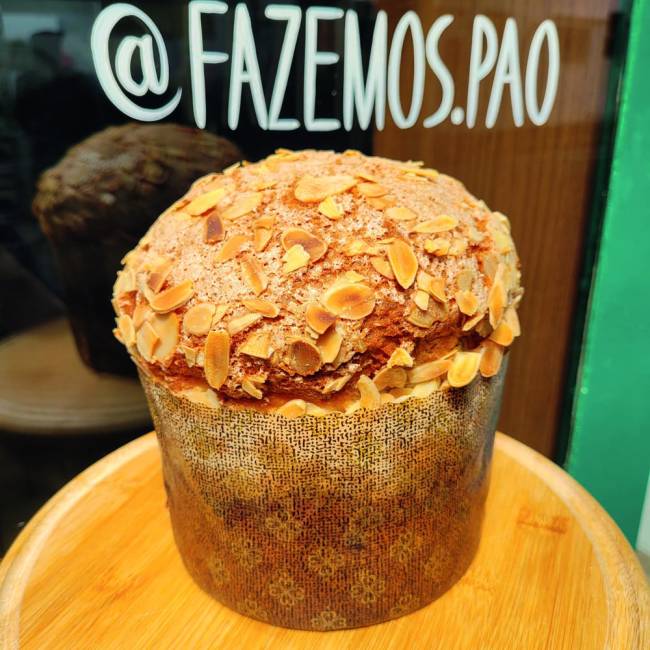 Fazemos Pão Panetone Panetone sobre mesa redonda de madeira clara com amêndoas açucaradas na casca