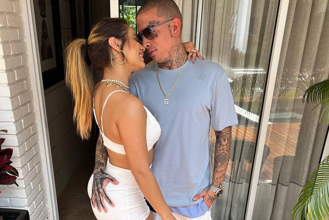 Foto de Lexa e Guimê em varanda de apartamento. Ela usa top e shorts branco e ele camiseta azul e shorts branco. Eles posam abraçados, se olhando