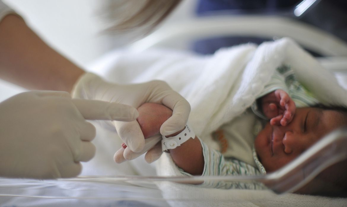 Foto mostra médico com luvas segurando mão de bebê recém-nascido