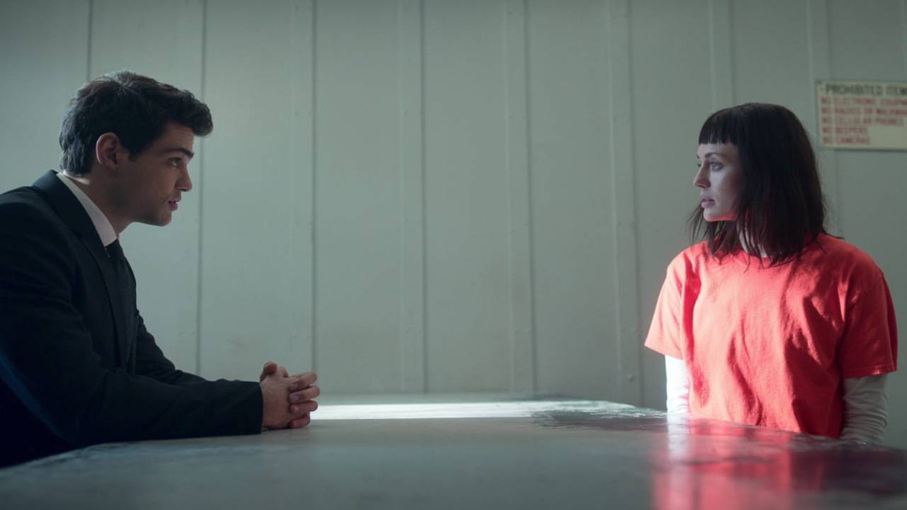 Jovem de terno e mulher prisioneira conversam em sala da prisão