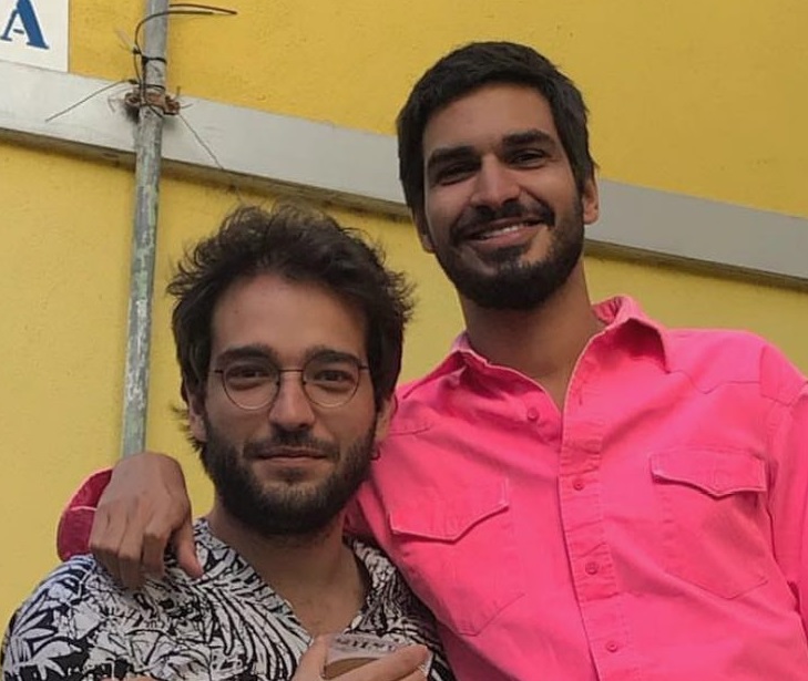 Humberto Carrão e Thales Junqueira abraçados sorrindo em frente a parede amarela