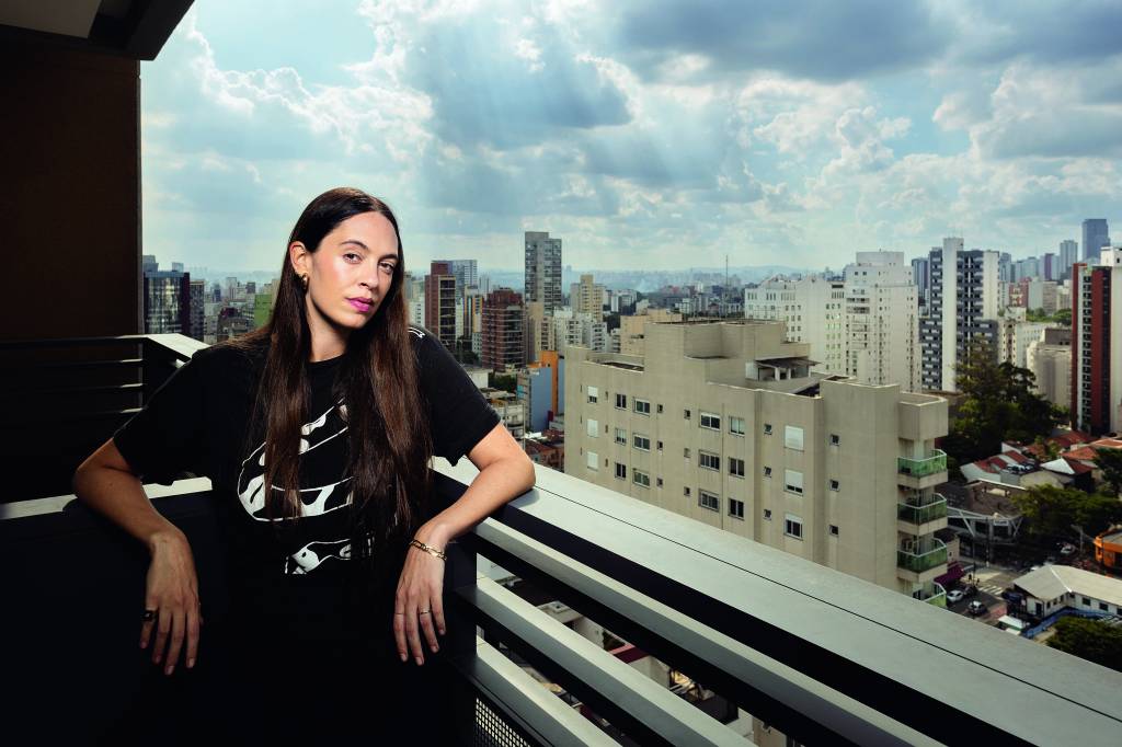 Imagem mostra mulher de roupa preta apoiada em sacada de varanda, com horizonte de prédios ao fundo