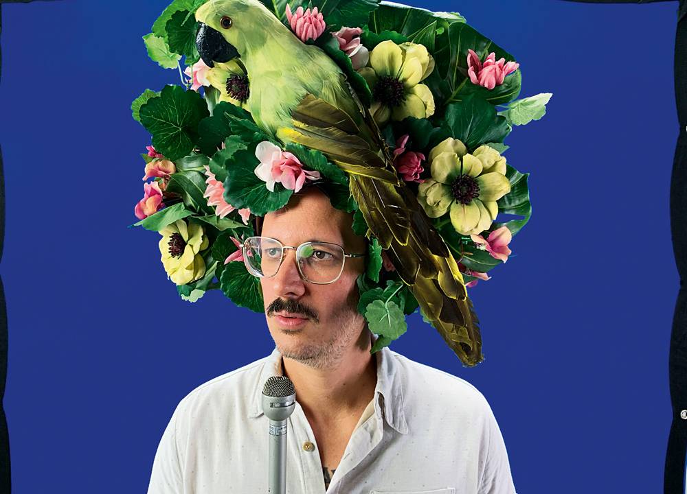 Imagem mostra homem de óculos e bigode com chapéu com flores e papagaio verde