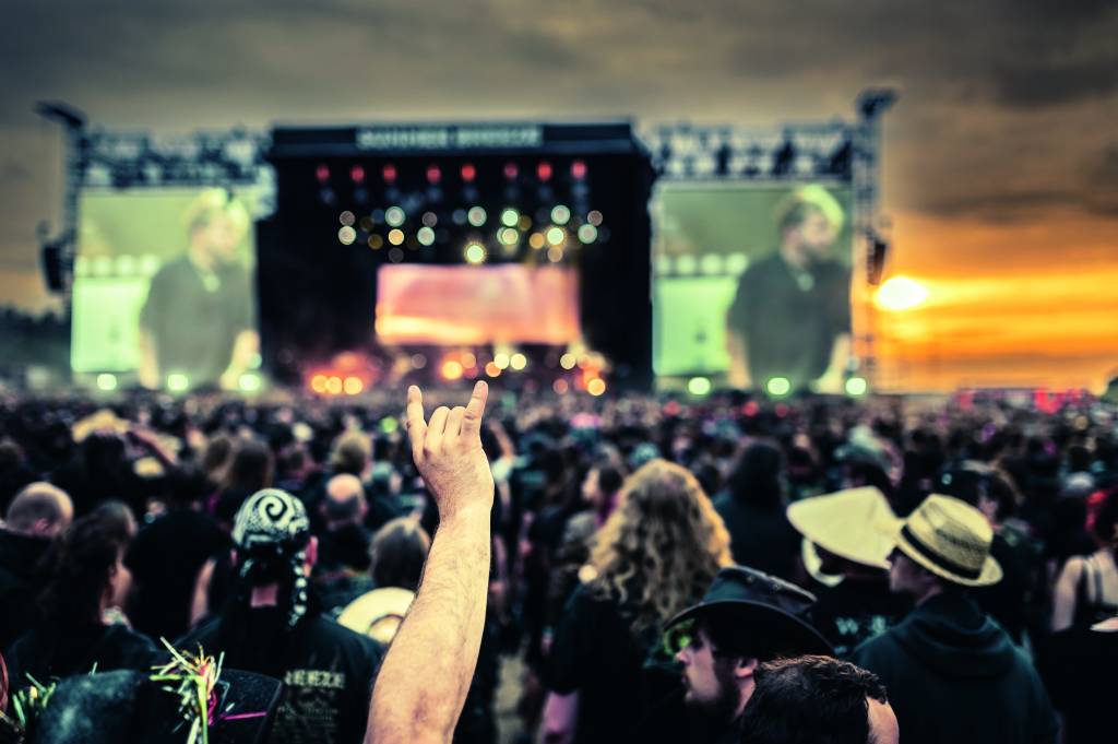 Imagem mostra mão fazendo sinal de rock'and'roll em meio a multidão, Ao fundo, desfocado, um palco