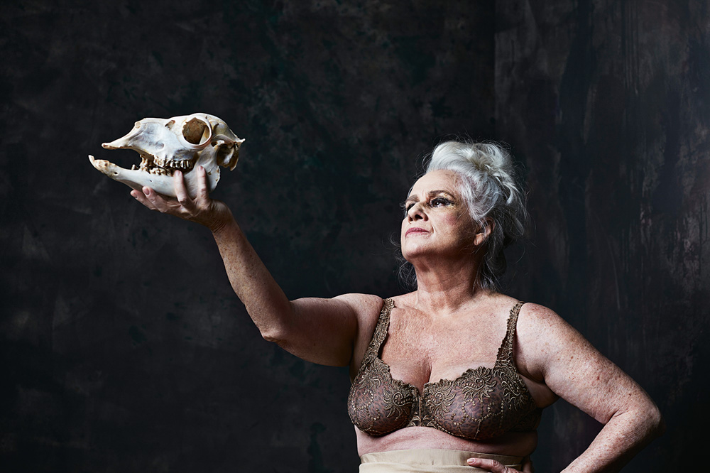 A atriz Vera Holtz, uma senhora idosa, branca, de cabelos lisos brancos presos em um coque, vista do busto para cima. Ela usa um sutiã marrom e segura no alto um crânio de animal