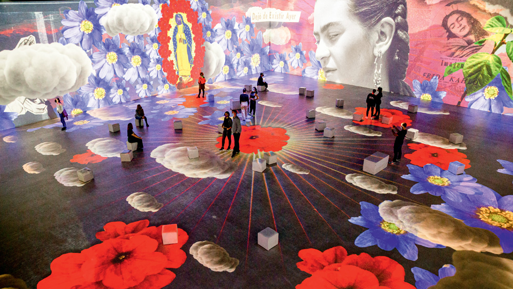 Frida Kahlo: A Vida de Um Ícone Grande salão com projeções de flores, itens da cultura mexicana e o rosto de Frida Kahlo