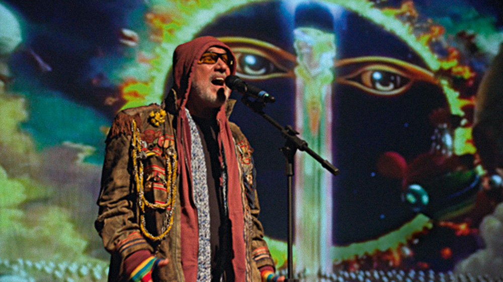 Cantor Zé Alexanddre, um homem idoso branco de barba grisalha, canta sozinho no palco. Usa uma jaqueta marrom com capuz e óculos de sol
