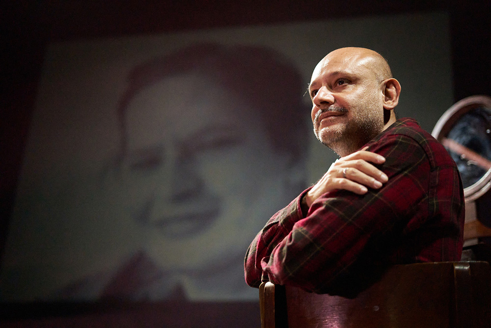 Nilton Bicudo, ator branco, magro, careca e de barba grisalha, em primeiro plano sentado em uma cadeira no palco. Ao fundo, uma tela exibe uma foto em preto e branco da poetisa Orides Fontela