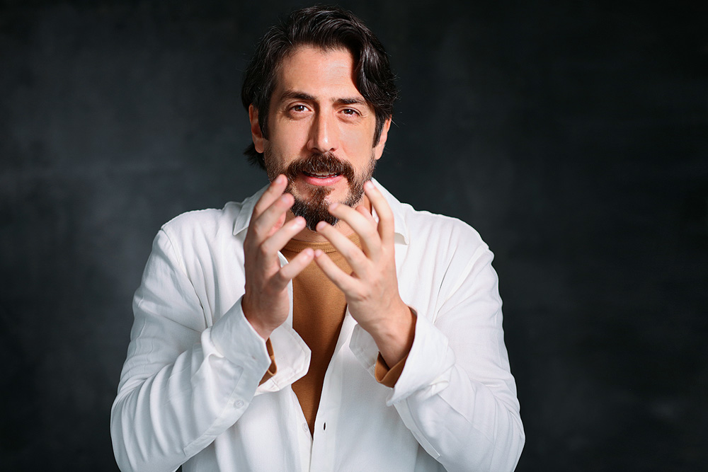 boa noite boa vista O ator Eduardo Mossri, um homem branco, jovem, de cabelos e barba pretos. Usa uma camisa branca aberta no peito e gesticula olhando para a câmera