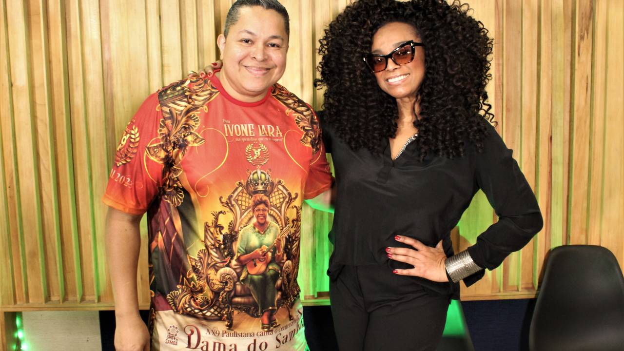 Darlan Alves e Paula Lima posam lado a lado, ele com camiseta laranja colorida, ela vestida de preto e óculos escuros.