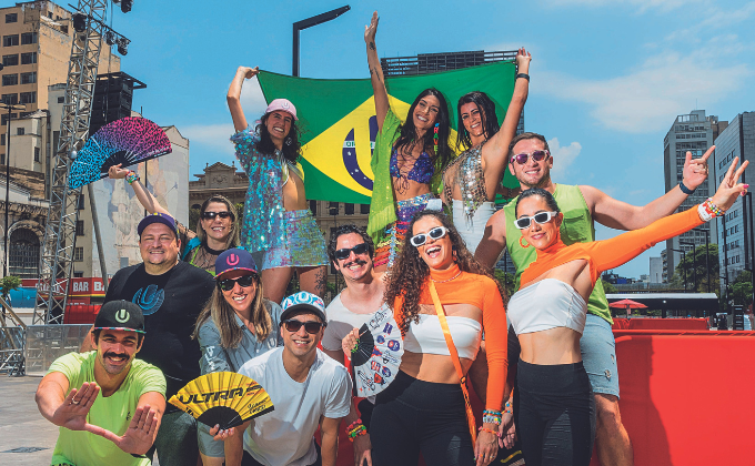 Imagem mostra grupo de pessoas vestindo roupas de carnaval, alegres, e segurando bandeiras e cartazes de festival de música. Ao fundo, um céu azul.