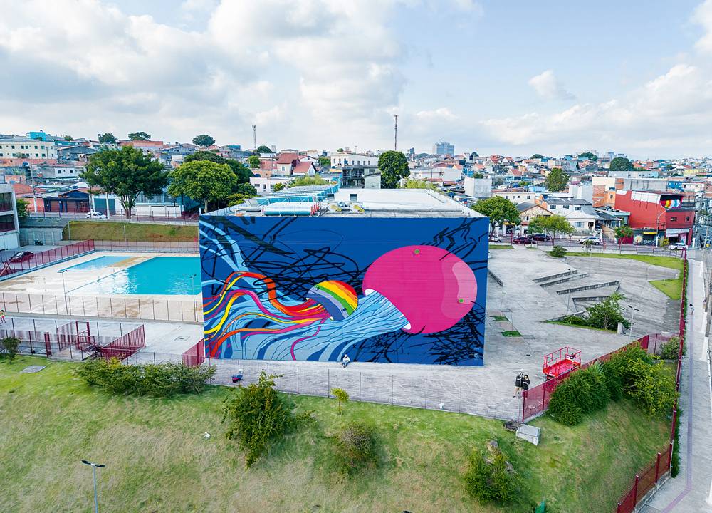 Foto mostra grafite em uma empena com uma criatura rosa com tentáculos azuis e uma criatura semelhante menor, colorida