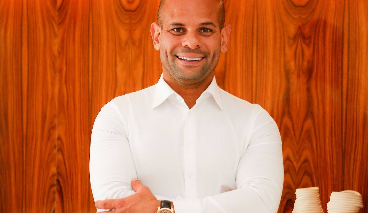 Homem negro e careca de braços cruzados sorri para a câmera. Veste camisa branca e relógio. Ao fundo, parede de madeira.