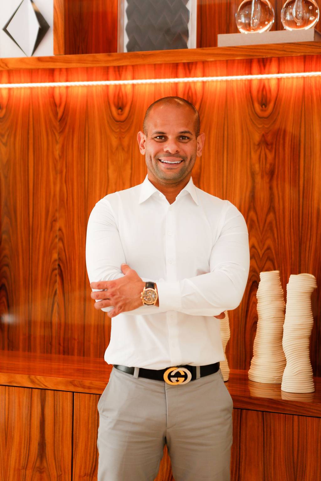 Homem negro e careca de braços cruzados sorri para a câmera. Veste camisa branca e relógio. Ao fundo, parede de madeira.