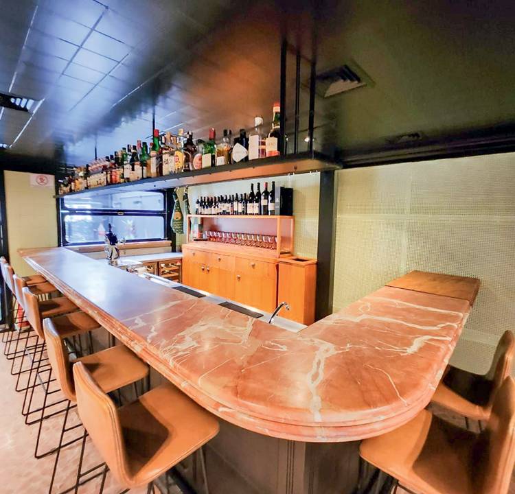 Ambiente interno de bar com balcão em mármore em formato de L e banquetas altas em metal e couro