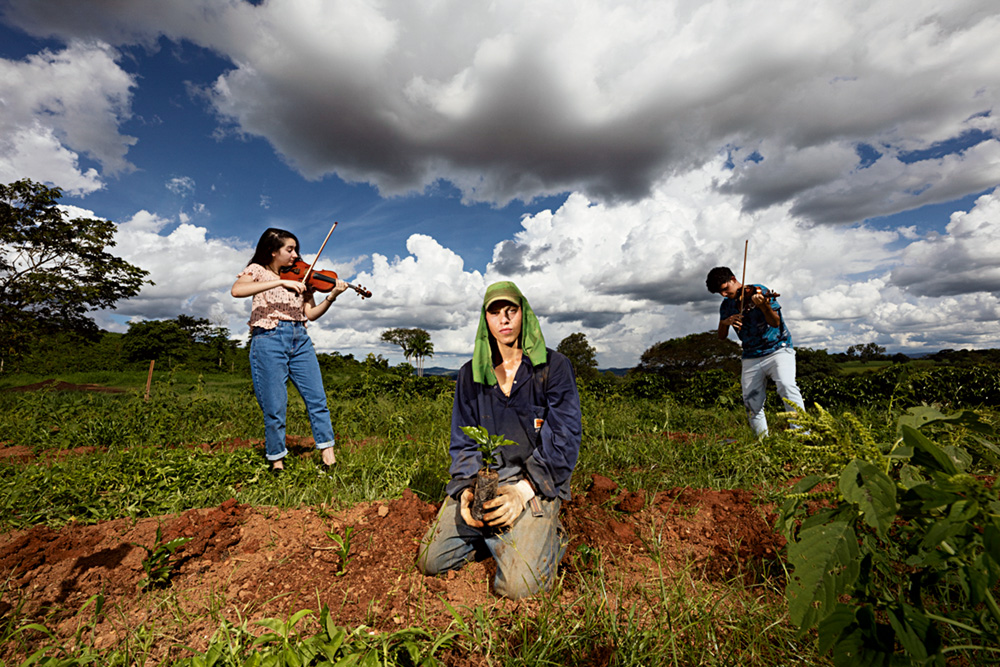 Imagem mostra três pessoas em gramado, um agricultor e dois violinistas