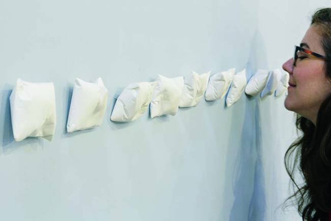 Diversos travesseiros de gesso fixados horizontalmente ao longo de uma parede cinza, ao fundo uma pessoa está cheirando um deles