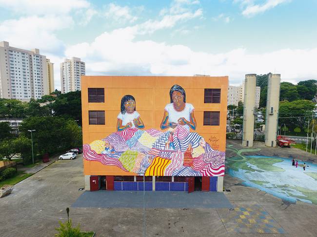 Foto mustra mural laranja com duas mulheres negras na lateral de prédio
