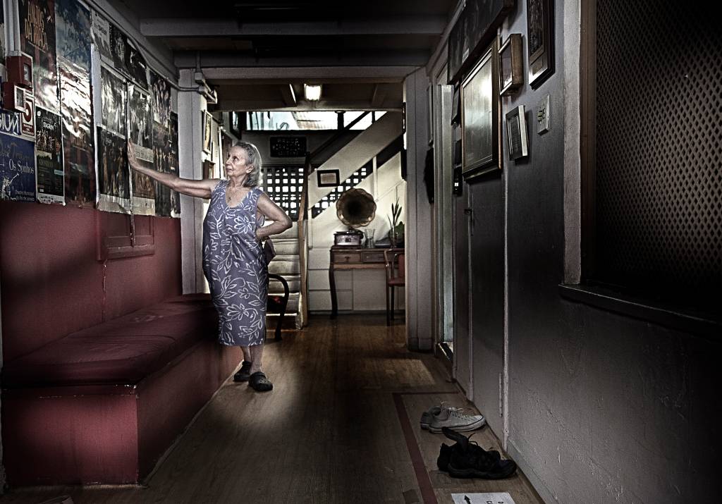 Imagem mostra mulher idosa apoiada em parede, usando vestido