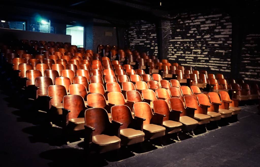 Foto exibe assentos de couro alinhados em sala de teatro iluminada.