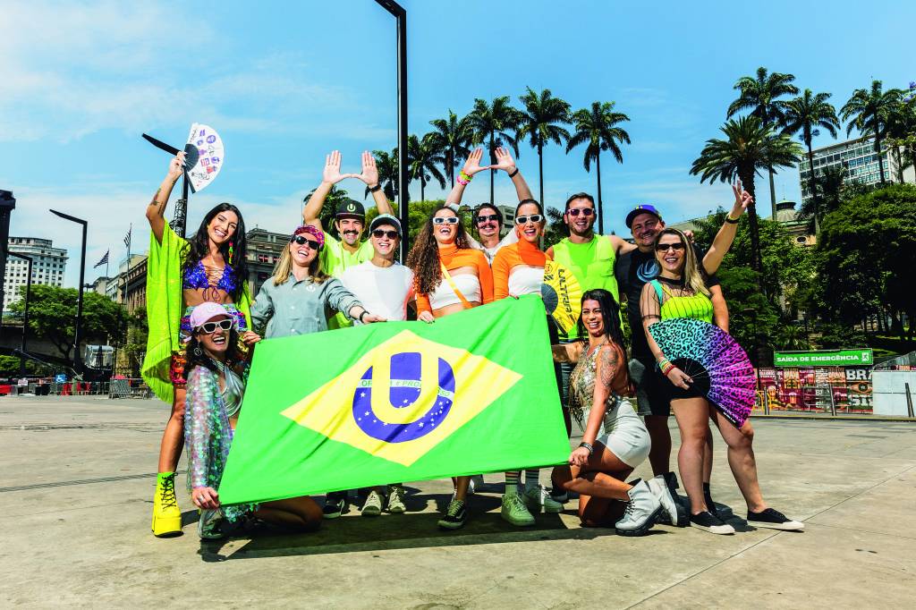 Imagem mostra grupo de pessoas vestindo roupas de carnaval, alegres, e segurando bandeiras e cartazes de festival de música. Ao fundo, um céu azul.