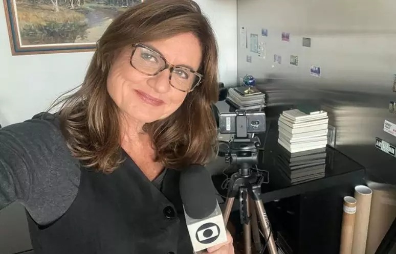 Selfie de mulher branca, com cabelo castanho, segurando microfone com logo da Globo, dentro de casa, com tripé e livros ao fundo