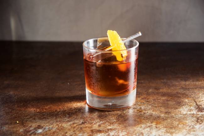 Copo baixo de vidro sobre mesa de madeira contendo líquido laranja escuro, gelo e fatia de picles de laranja espetada em palito no drink