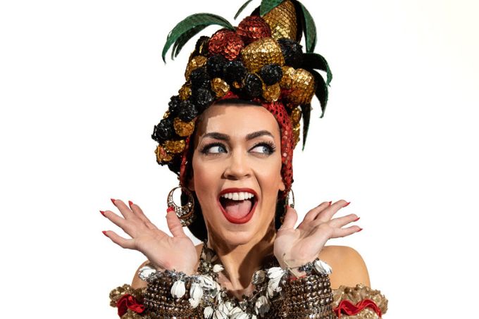 Laila Garin, atriz branca e magra, com figurino de Carmen Miranda. Ela sorri, olha para os lados e estende as mãos ao lado do rosto